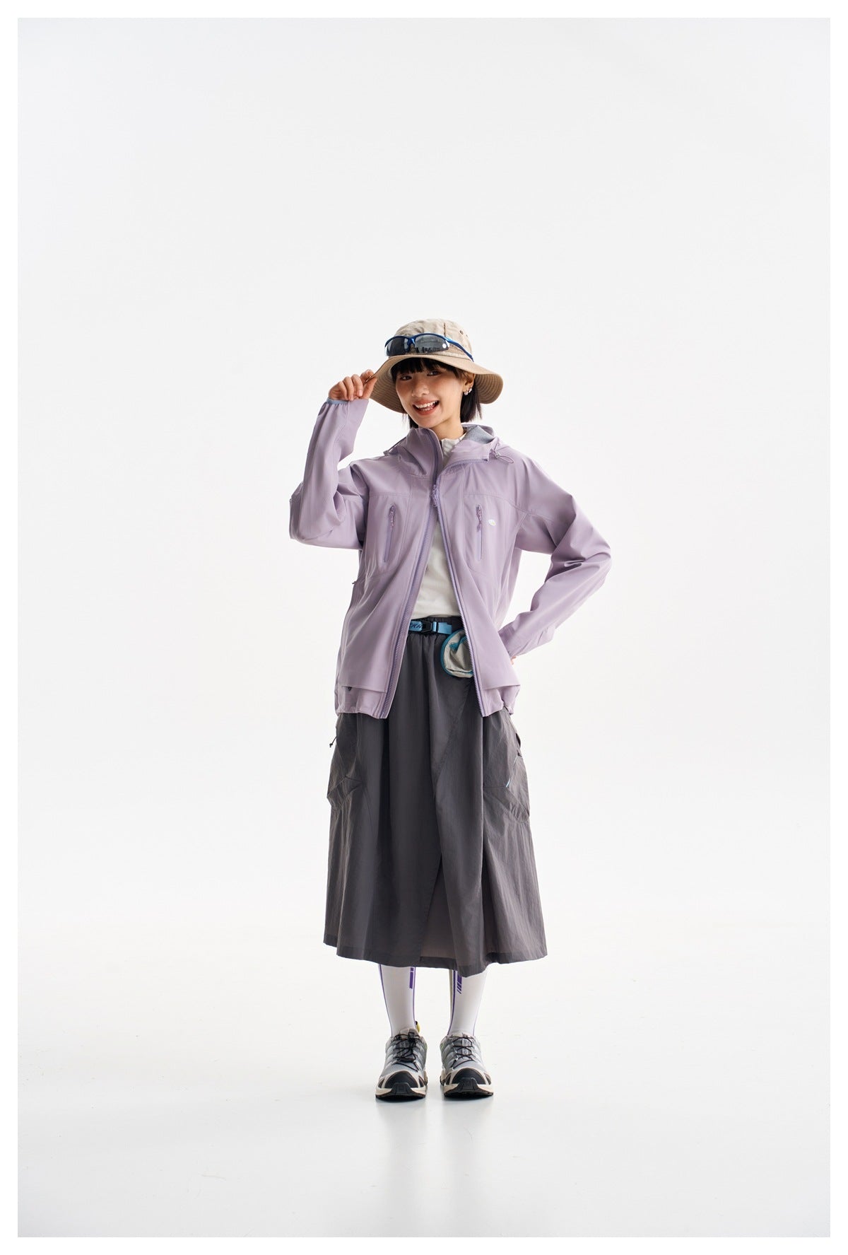 Model wearing a lavender hooded jacket with a dark long skirt and a beige sun hat.