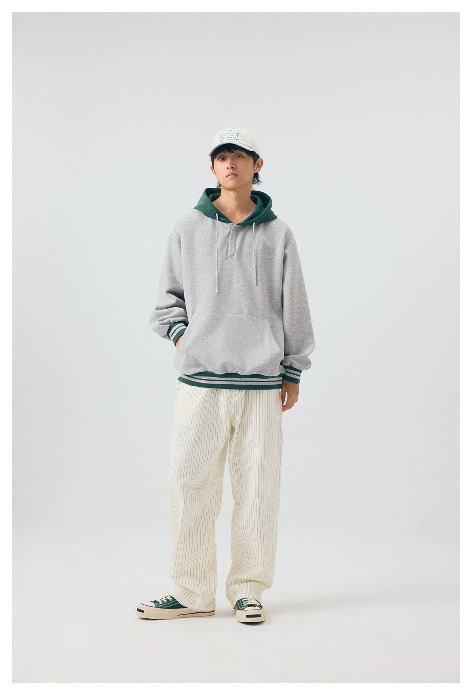Full-length front view of a model in a light gray hoodie with a dark green hood and striped trim.