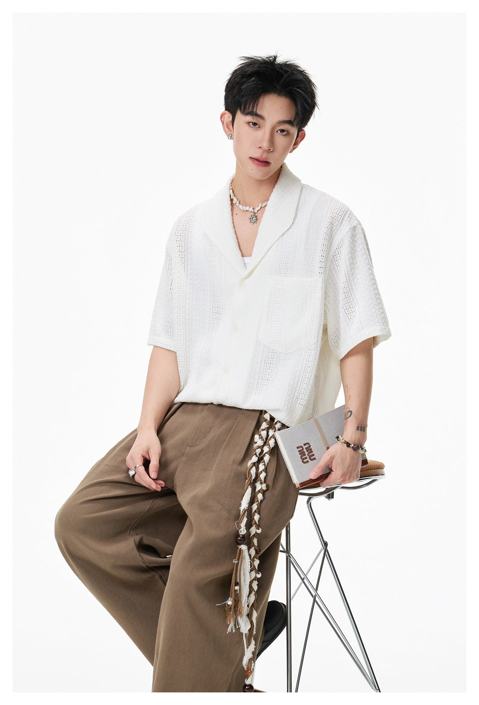 Seated model wearing a white openwork short-sleeve shirt with shawl collar and chest pocket, paired with brown trousers.