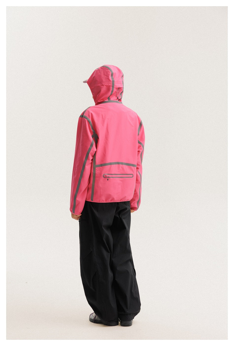 Back view of a model wearing a bright pink hooded jacket with gray trim and loose black trousers.