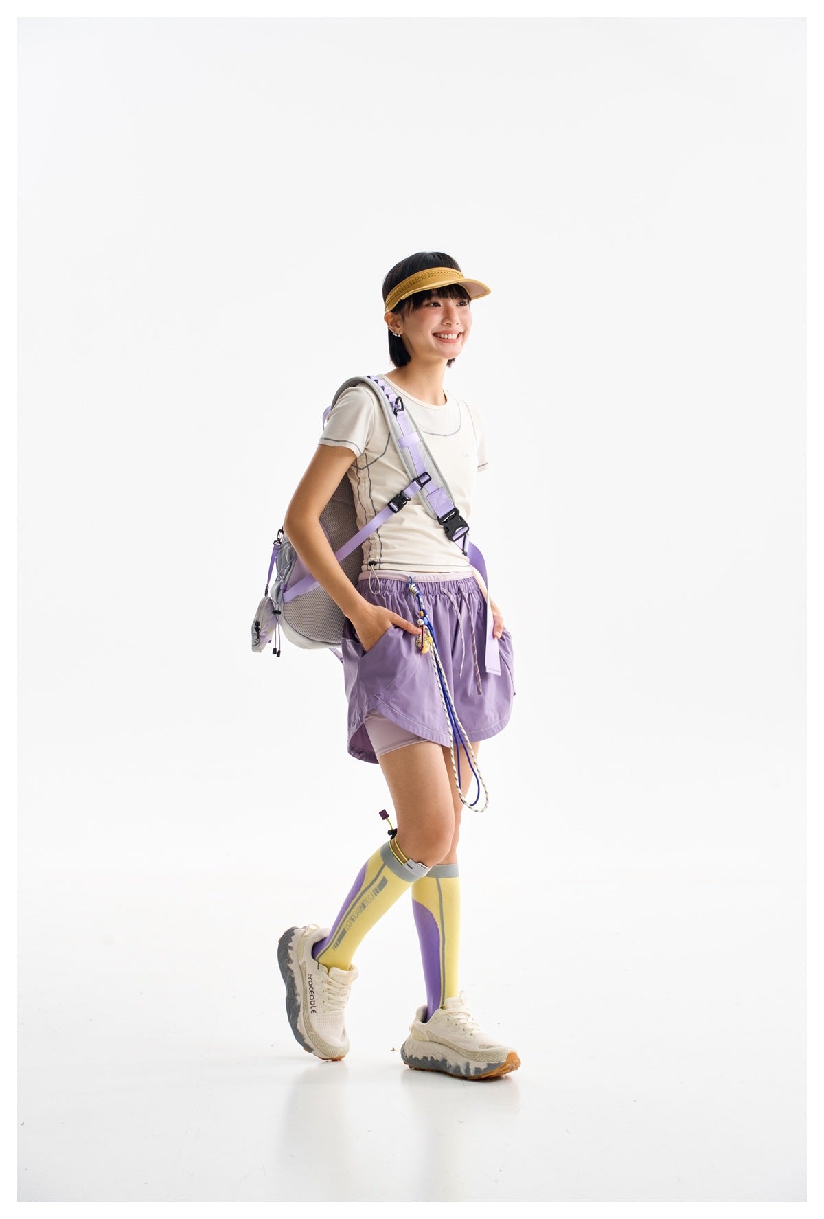 Model in profile wearing an ivory contrast-stitched tee, lavender shorts and a backpack with yellow-purple socks.