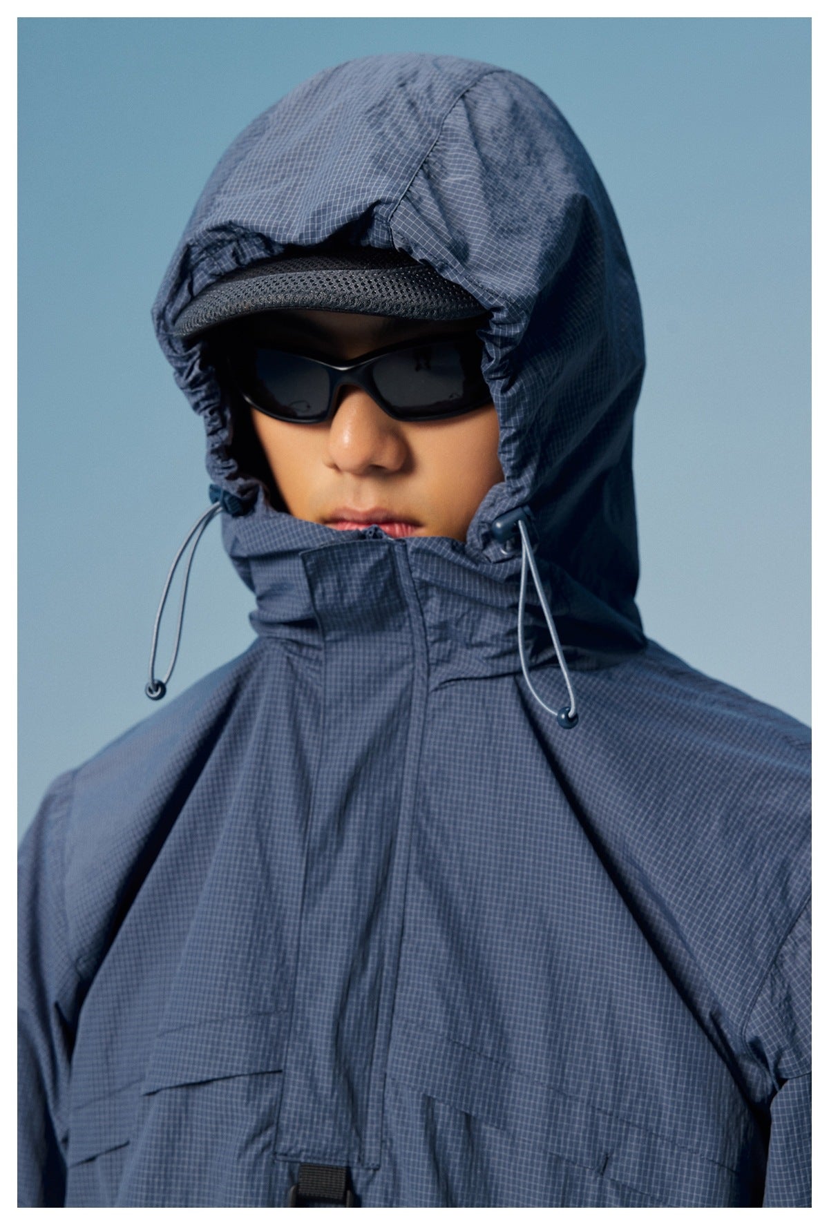Close-up of a model in a slate-blue grid-pattern anorak, showing the hood, drawcords and high collar.