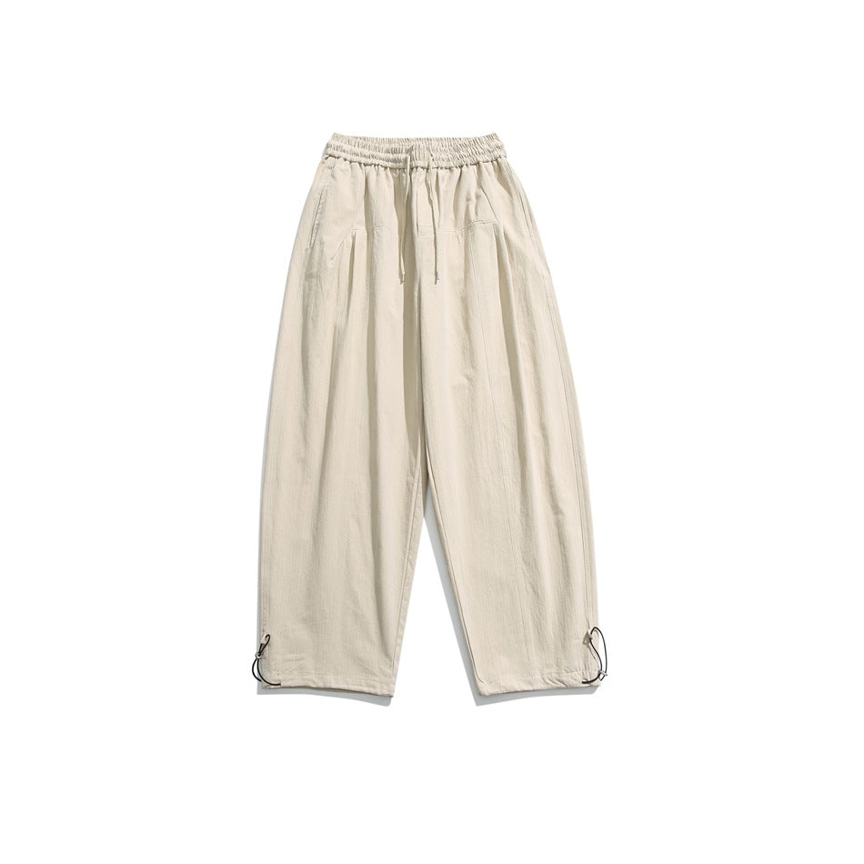 Beige wide-leg drawstring pants with elastic waistband, front pleats and toggle hem cinches, flat lay on white