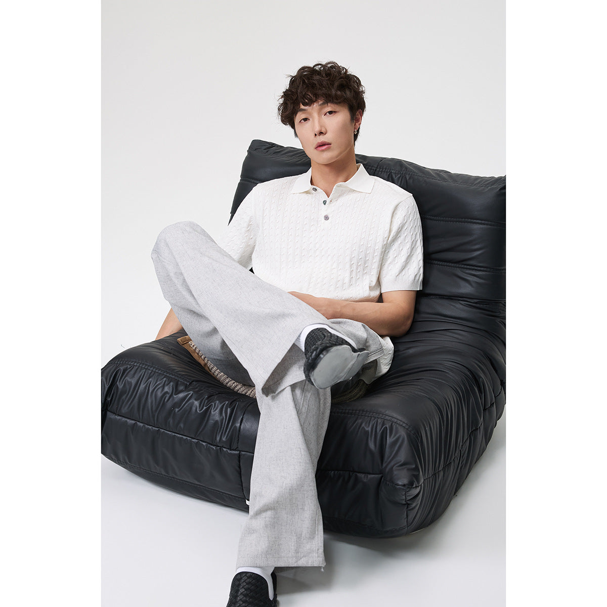 Model seated in a black chair wearing a white textured short-sleeve polo and light gray trousers.
