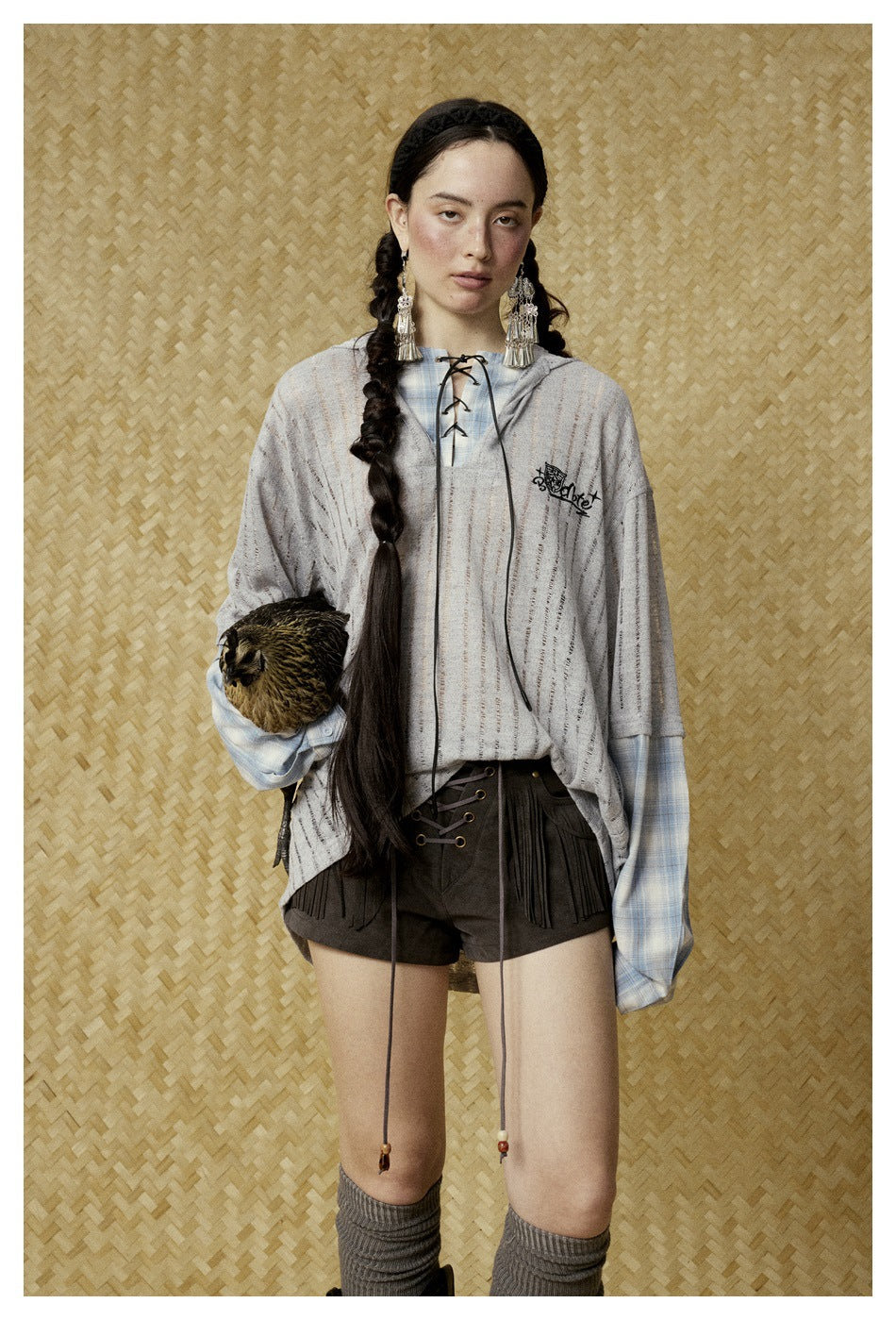 Model wearing gray distressed knit hooded top with lace-up neckline and plaid sleeves, with brown fringe shorts