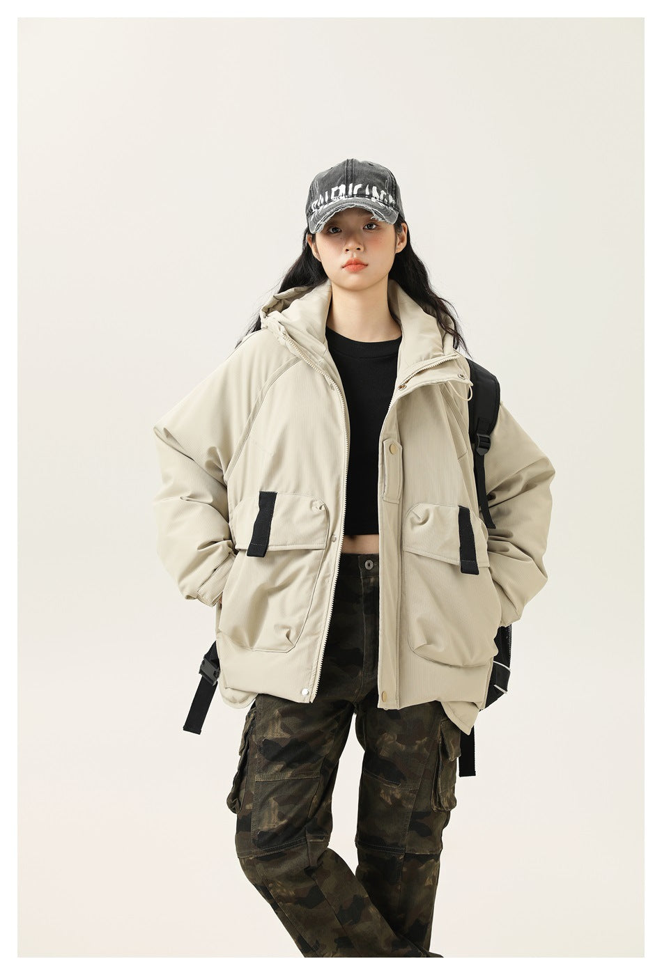 Model wearing an open light cream hooded jacket with large flap pockets and black accents, styled with camouflage pants.