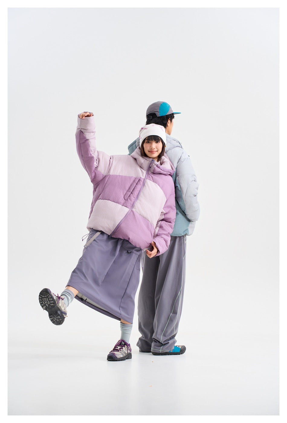 Two models pose together; the woman wears a color-block lavender and pale pink hooded puffer jacket, and the man wears a pale