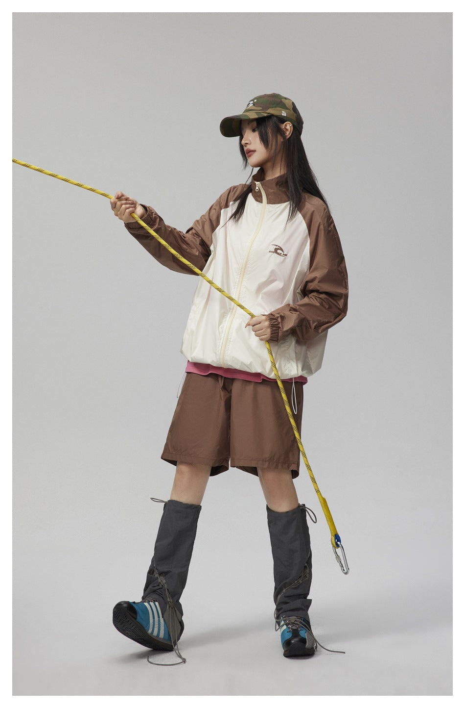 Model wearing a cream zip-front jacket with brown sleeves and brown shorts, with a camouflage cap and climbing rope.