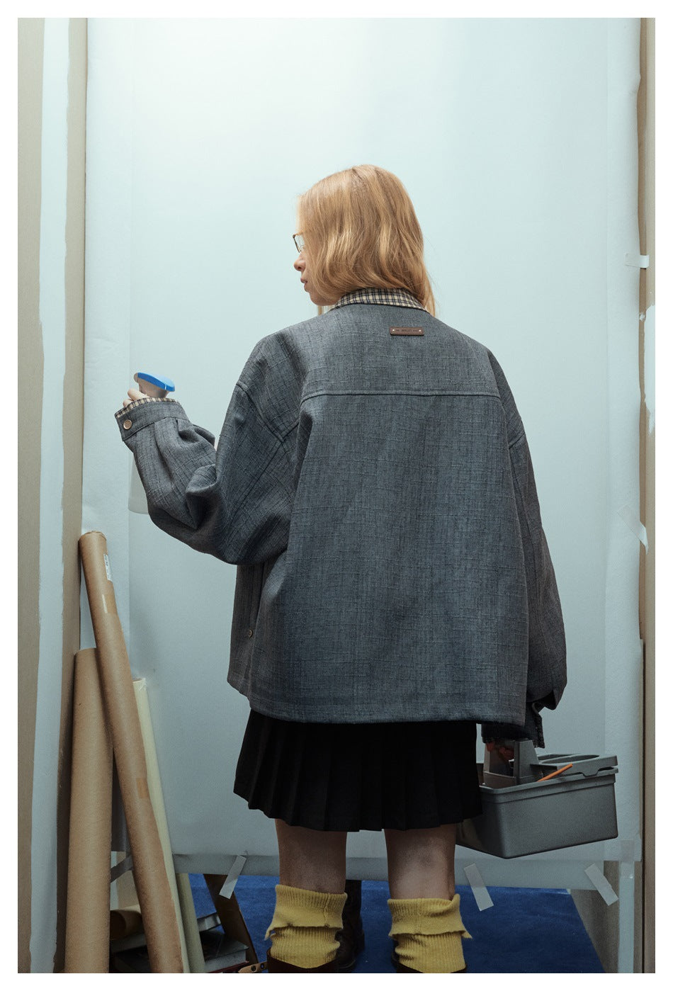 Back view of a model wearing an oversized charcoal jacket with a checked collar and black pleated skirt.