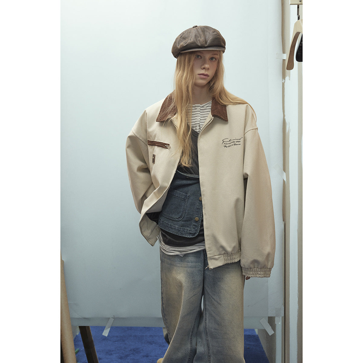 Model wearing a cream zip jacket with a brown collar, layered tops, faded wide-leg jeans, and a brown cap.