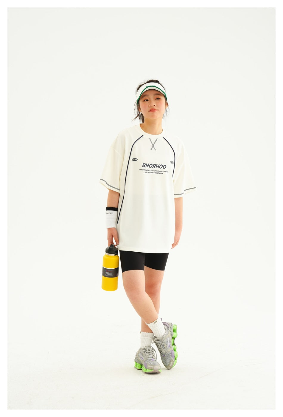 Model wearing an oversized ivory T-shirt with dark piping and chest print, black shorts, and a white-and-green visor.