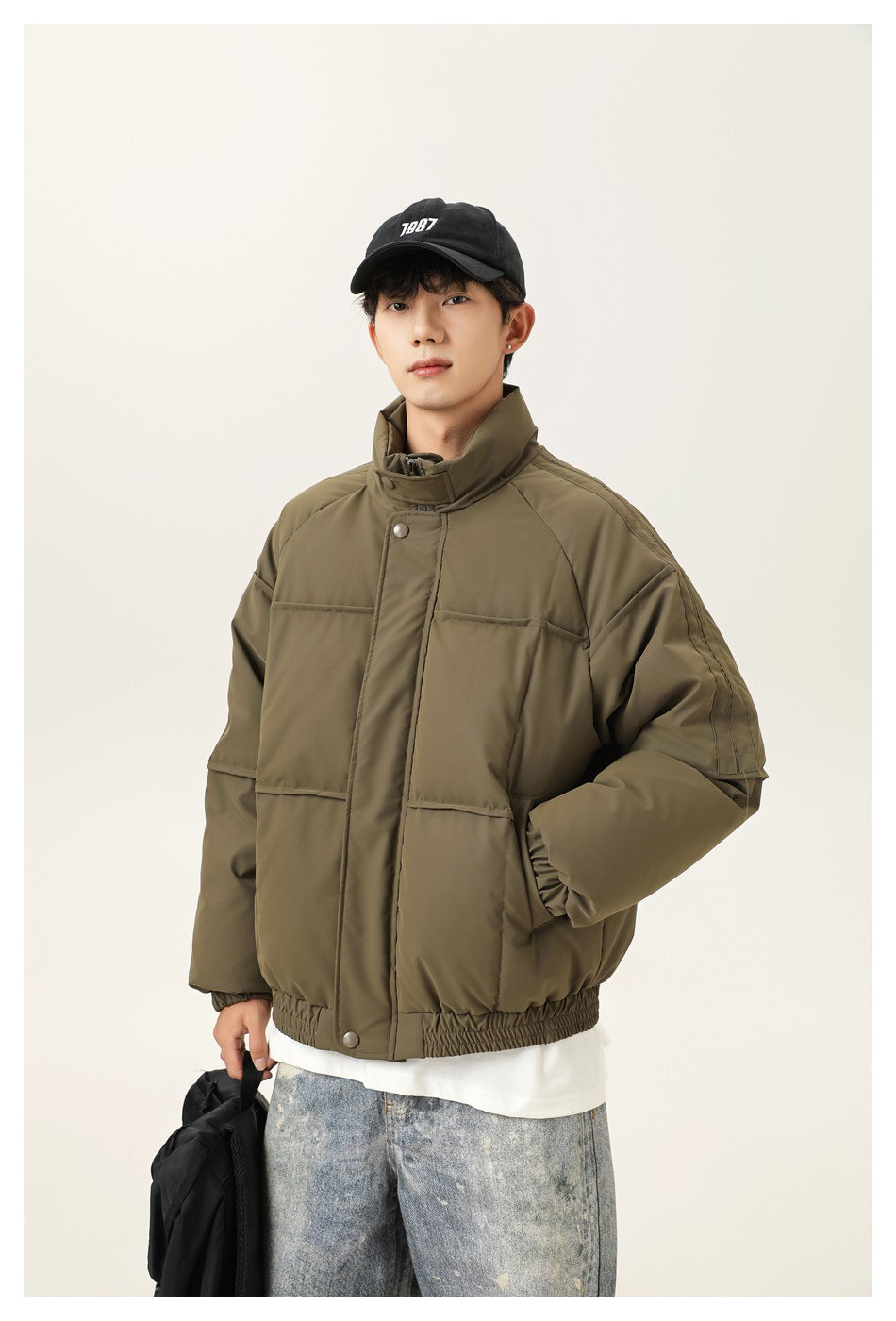 Man wearing an olive padded jacket with a high collar, snap placket, front pockets, and elastic hem; black cap has 1987