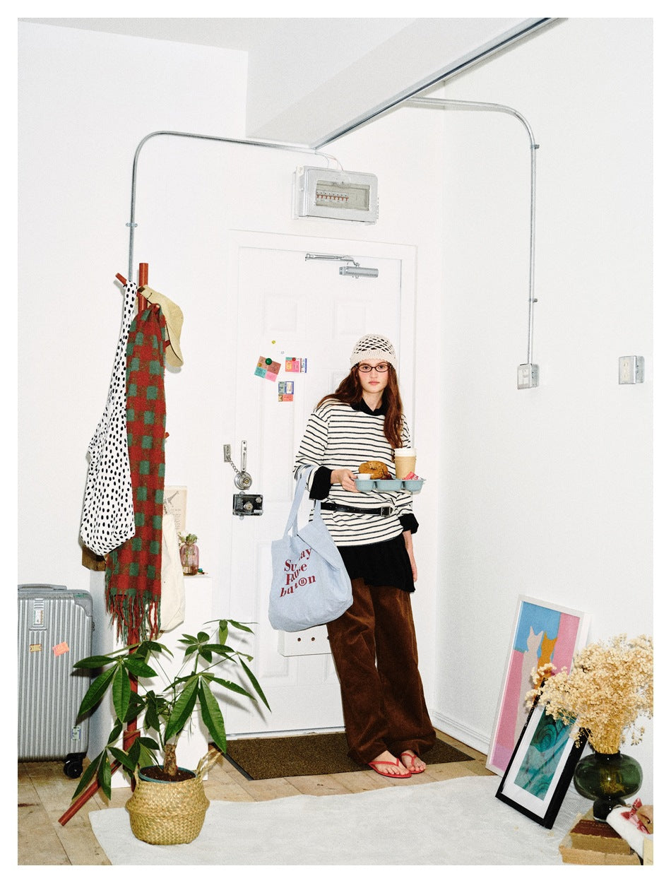 Model in a striped long-sleeve top with black layers and brown wide-leg trousers, standing by a door