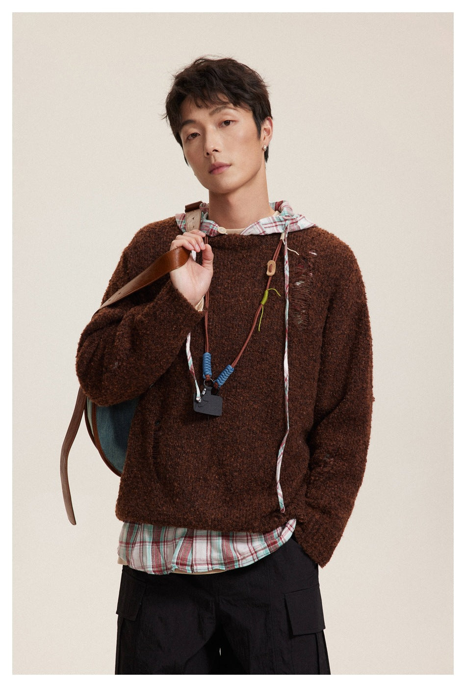 Model wearing a brown distressed crewneck sweater over a plaid hooded shirt and black cargo trousers.