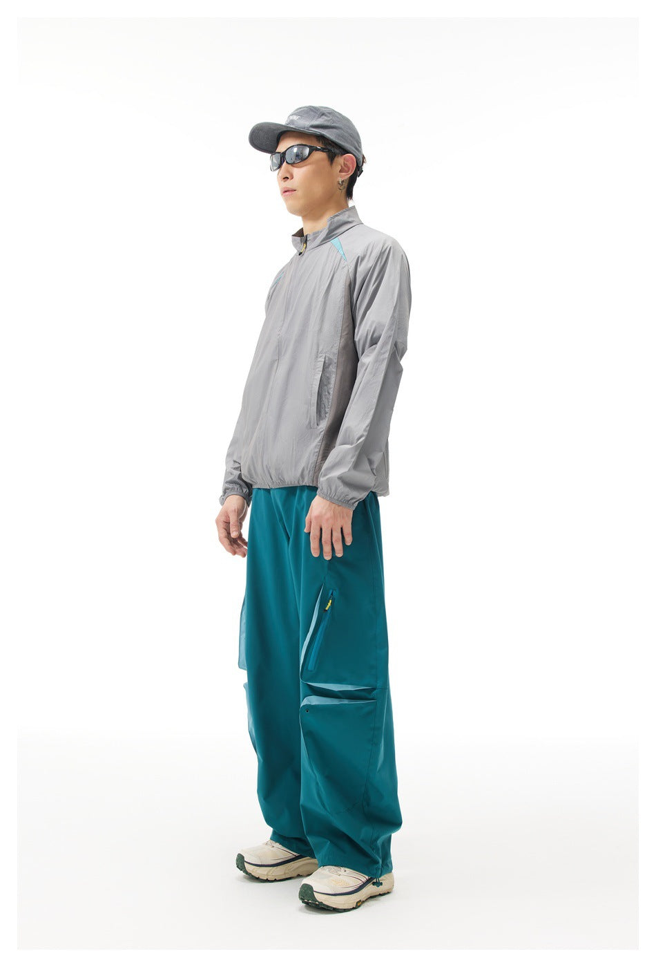 Side view of a man wearing a light gray zip jacket with turquoise shoulder accents and teal cargo trousers.