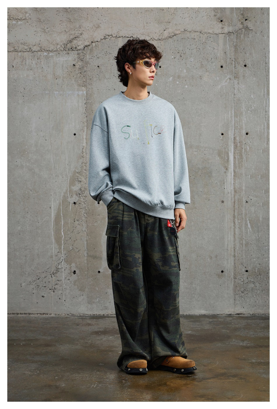 Full-length view of a model in a gray oversized crew-neck sweatshirt with camouflage cargo pants and tan clogs