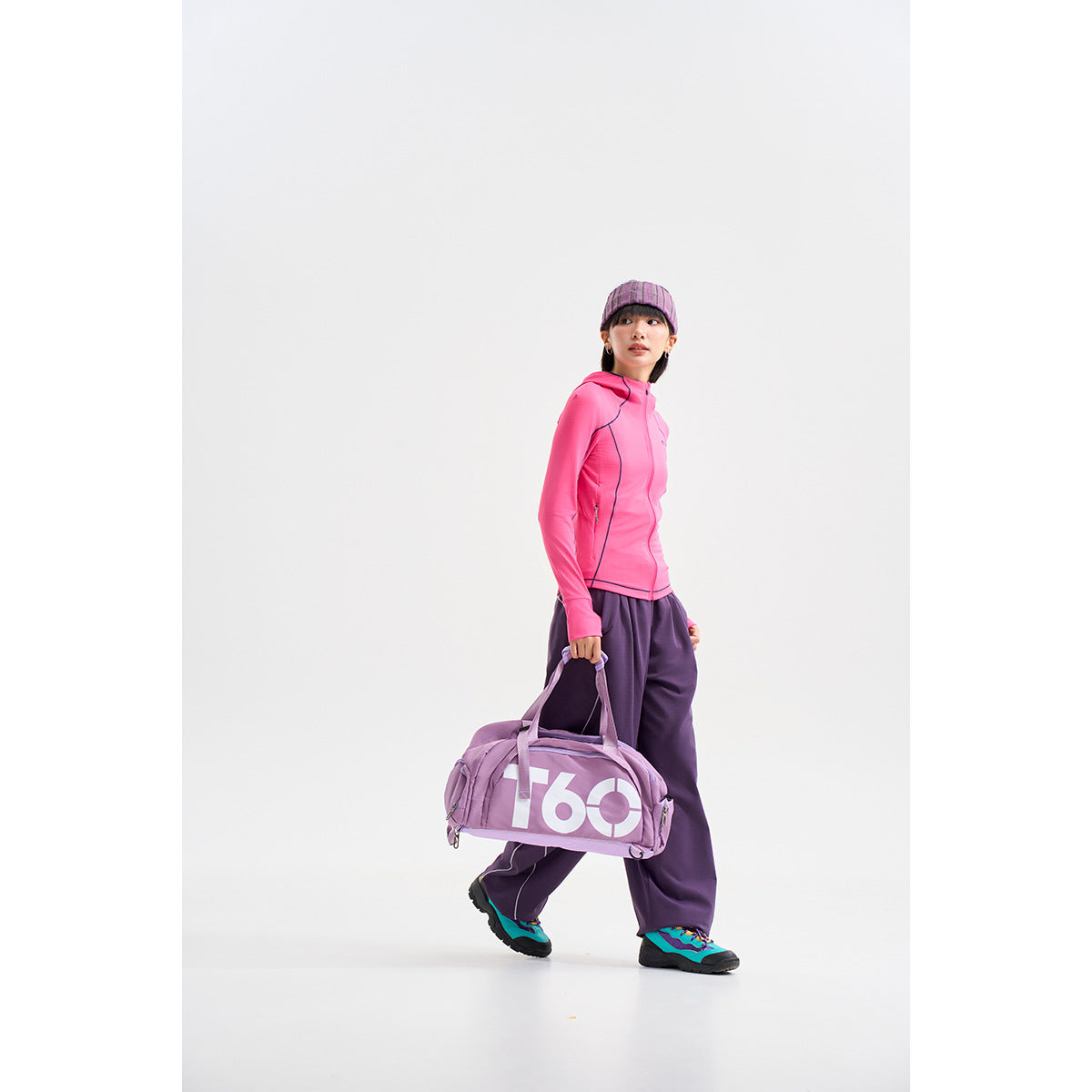 Model wearing a bright pink zip-up hooded jacket and loose purple pants, carrying a lilac duffel bag.