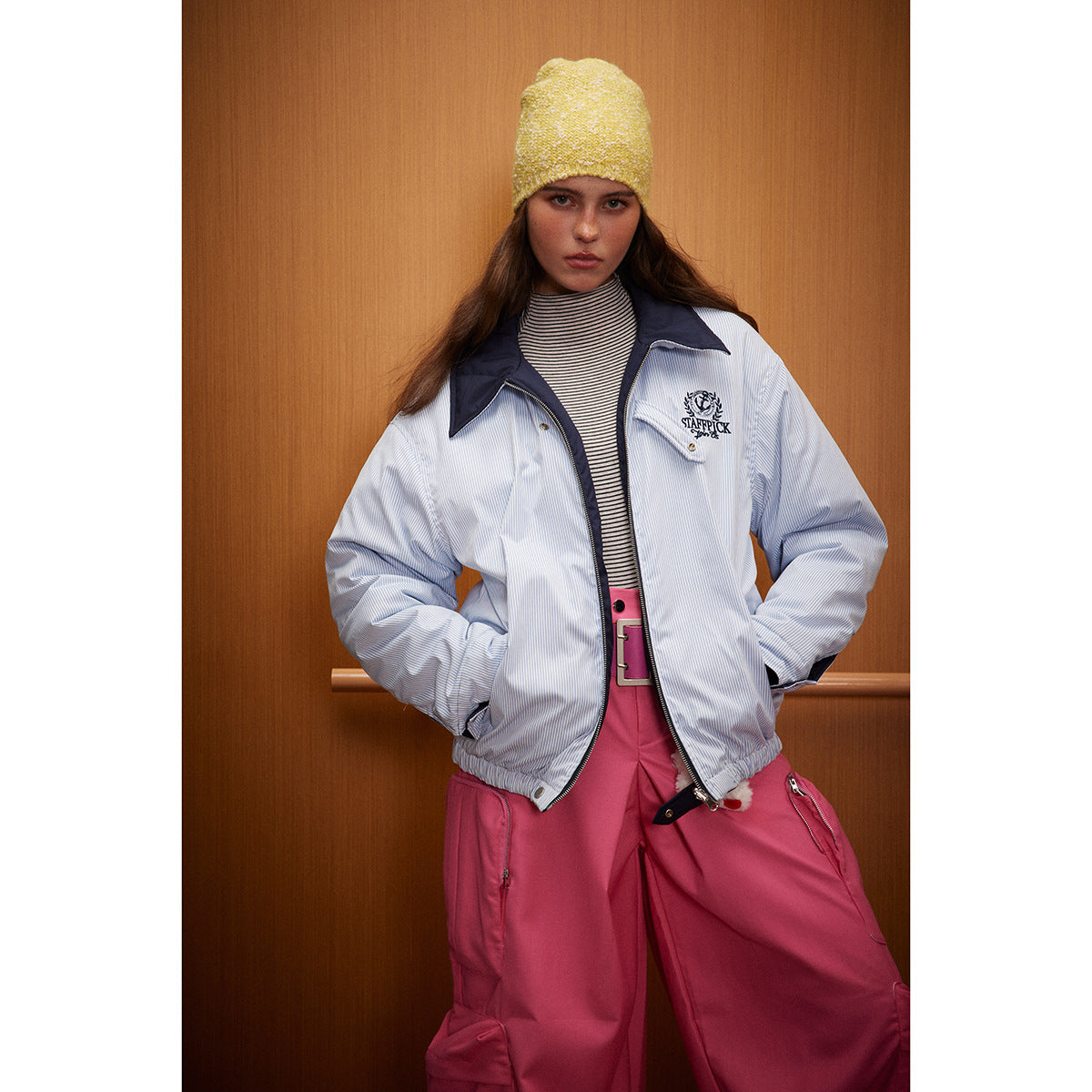 Model wearing a light blue pinstriped zip jacket with a navy collar, embroidered crest and pink trousers.