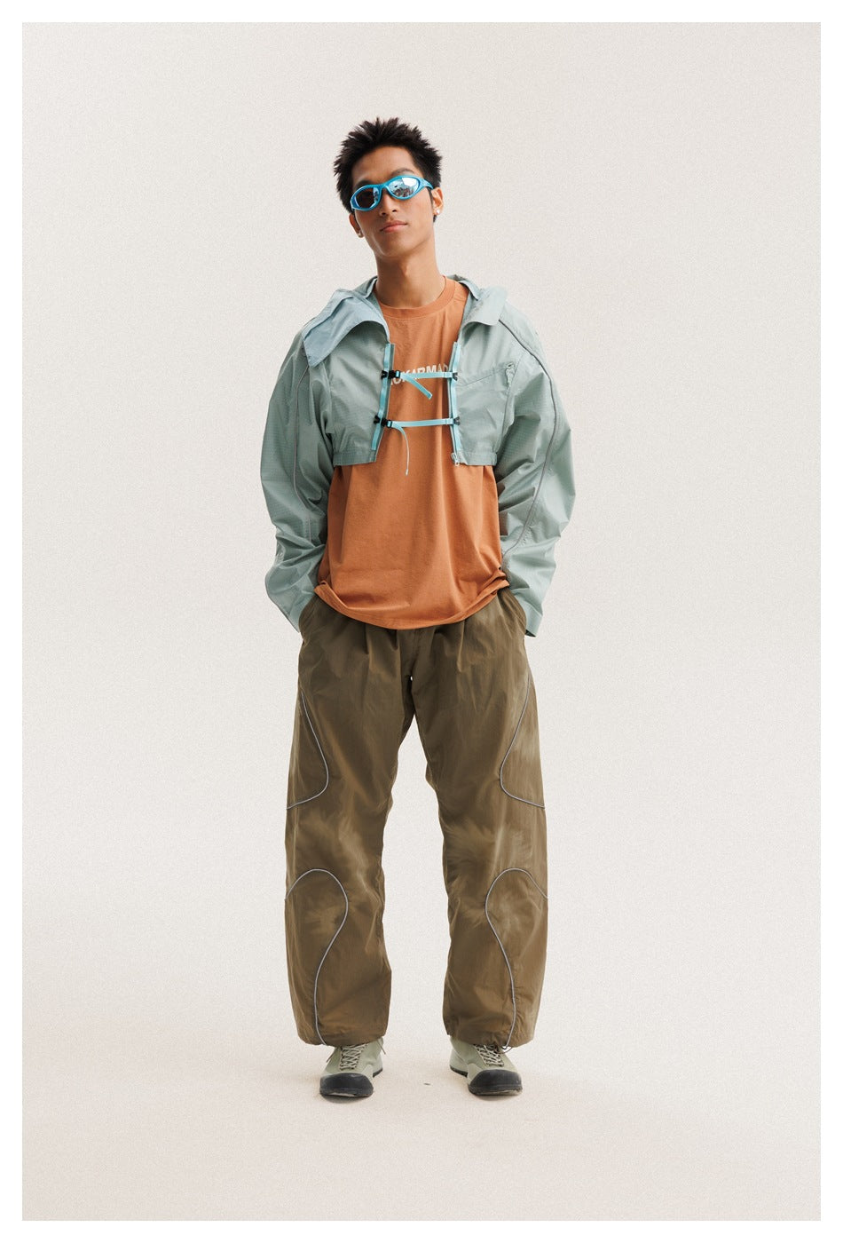 Model wearing a pale sage jacket open over a rust-orange T-shirt, with loose brown pants and blue sunglasses.