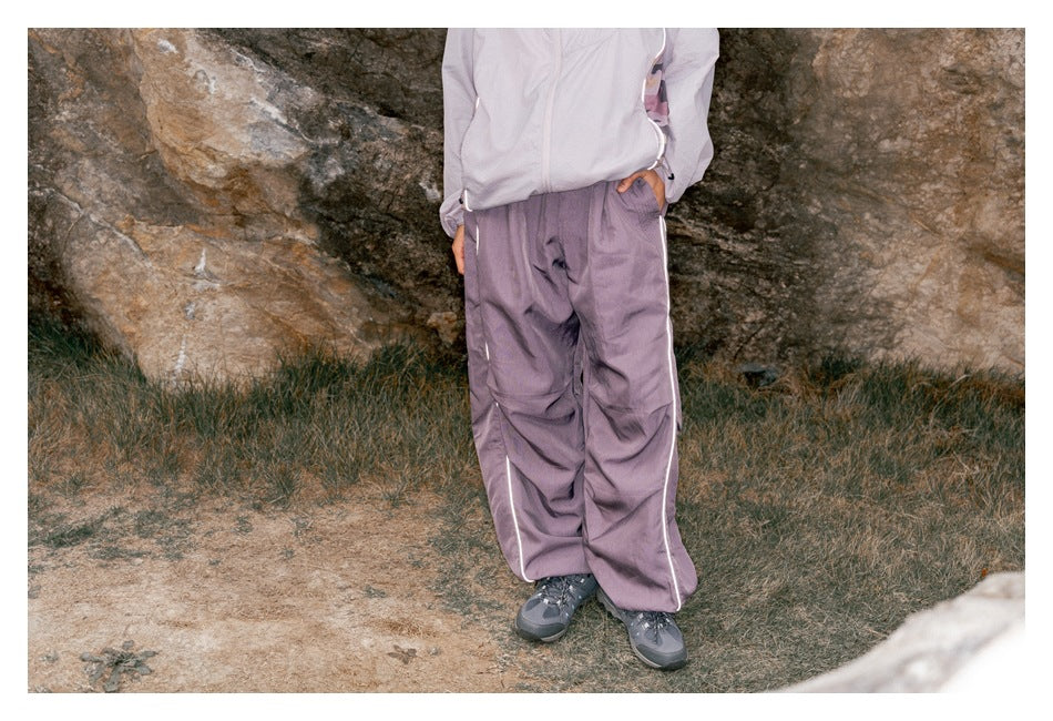 Front view of a model wearing loose muted lavender pants with white side piping outdoors.