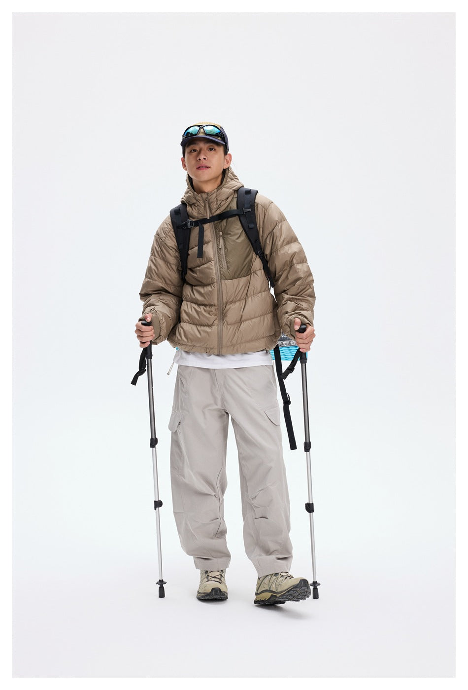 Front view of a model wearing a taupe hooded puffer jacket and light gray trousers with a backpack and trekking poles.