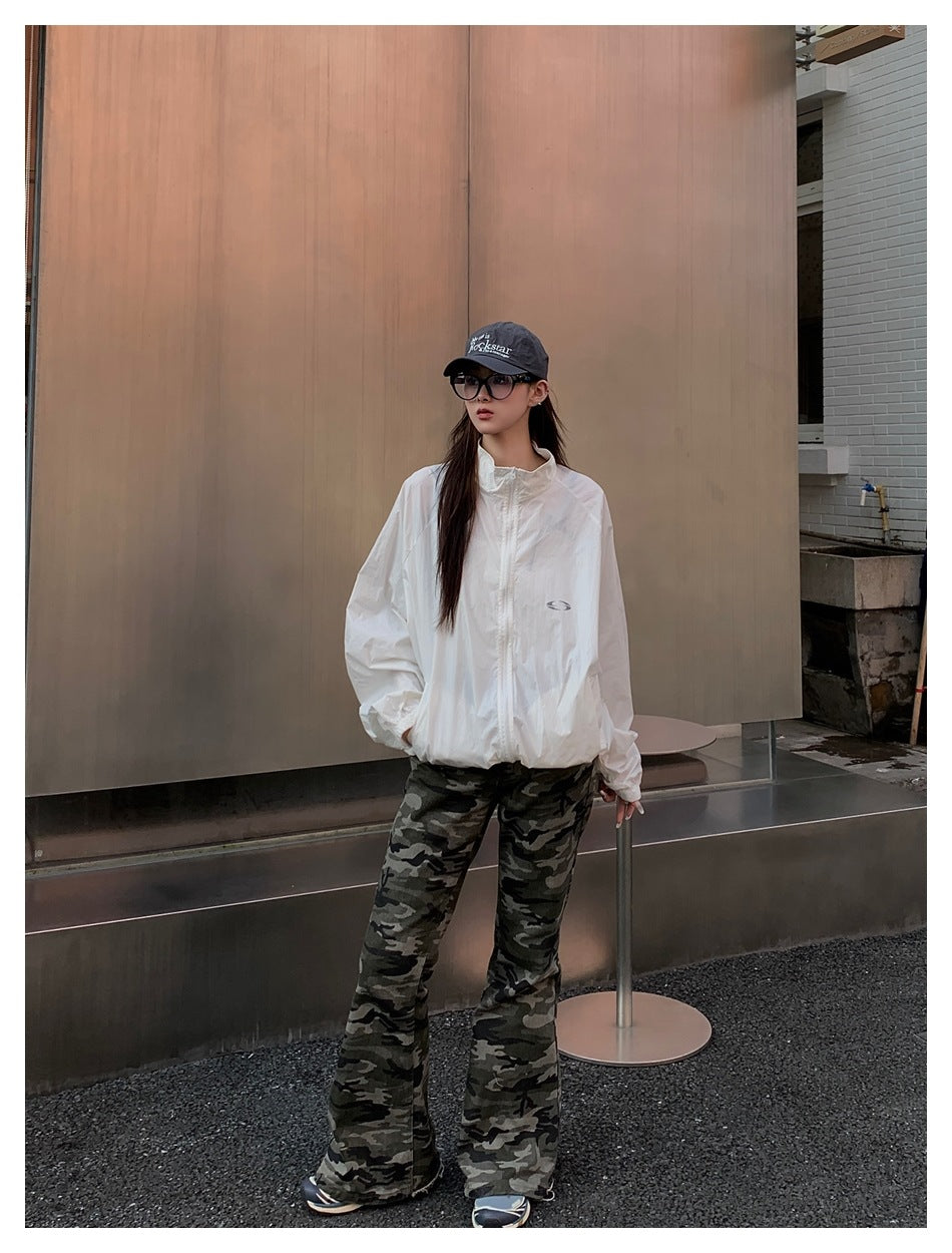 Model in an oversized white zip jacket with camouflage flared pants and a dark cap in front of a metal wall