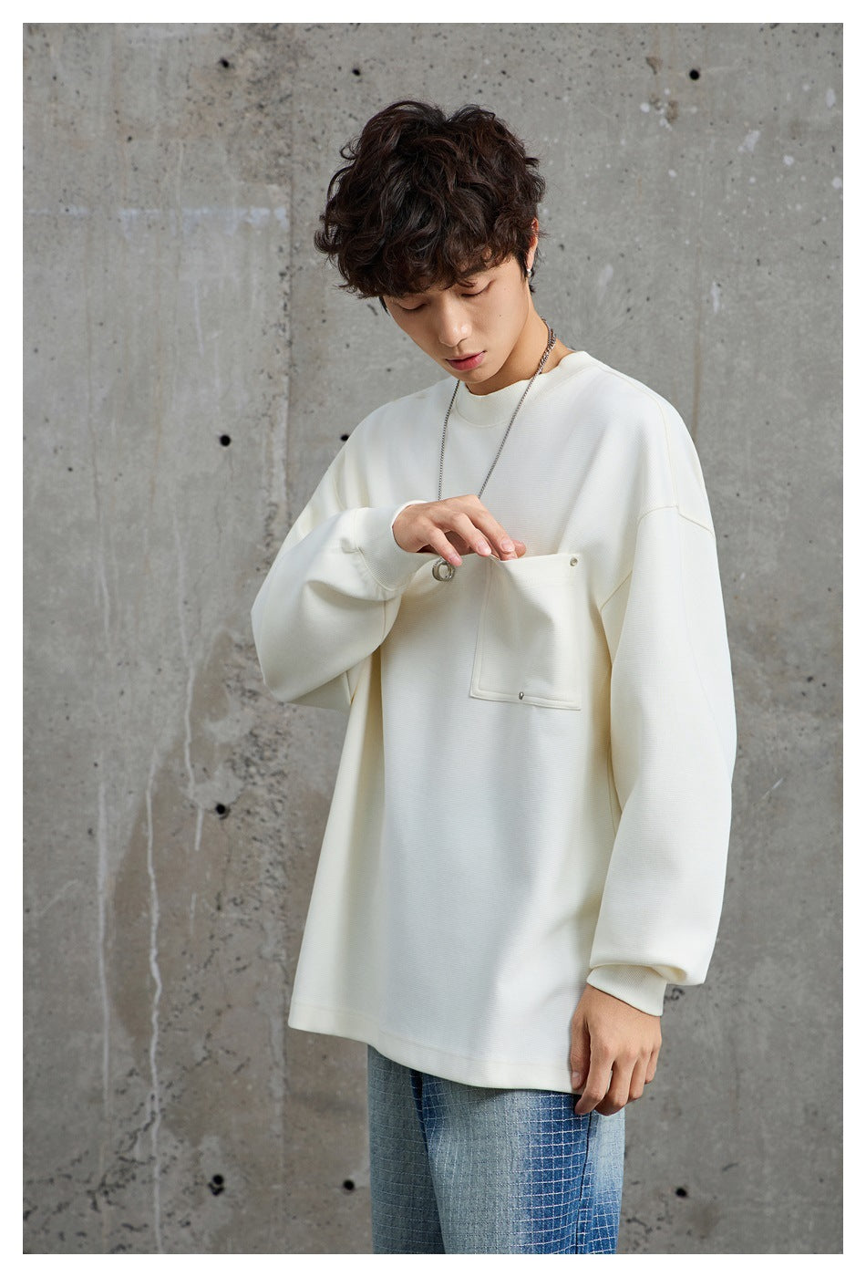 Model reaches into the large chest pocket of an oversized ivory sweatshirt, showing its pocket hardware and relaxed fit.