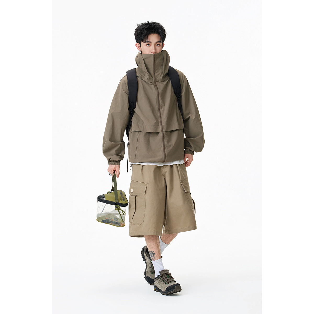 Model wearing a taupe high-collar zip jacket and matching cargo shorts, with a backpack and outdoor gear.