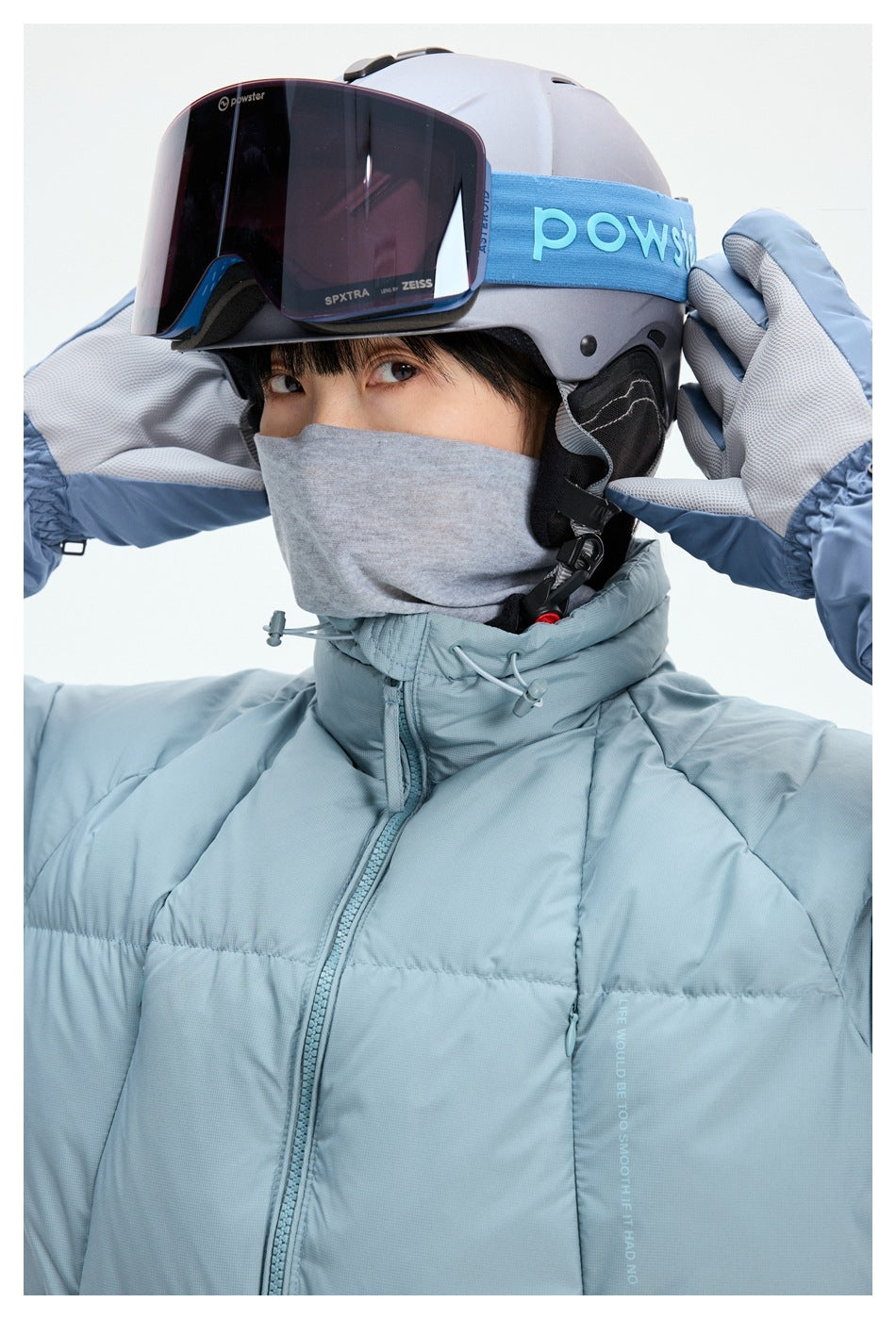 Close-up of a pale blue puffer jacket's high collar and zipper, worn with ski goggles, helmet, and gloves.