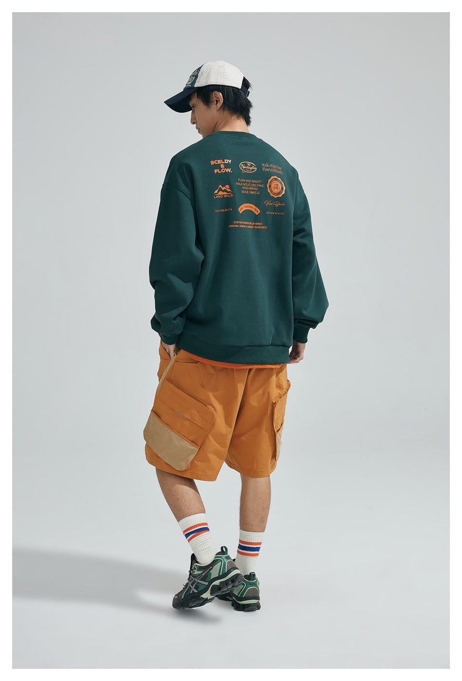 Model seen from the back wearing a dark green sweatshirt with orange back prints, orange cargo shorts and cap