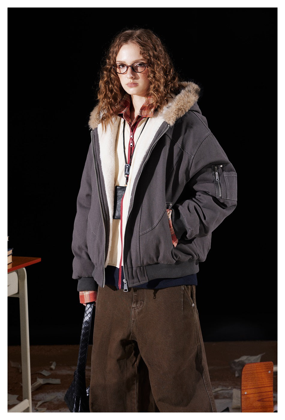 Model wearing open dark gray hooded jacket with cream fleece-like lining over a plaid shirt and brown pants