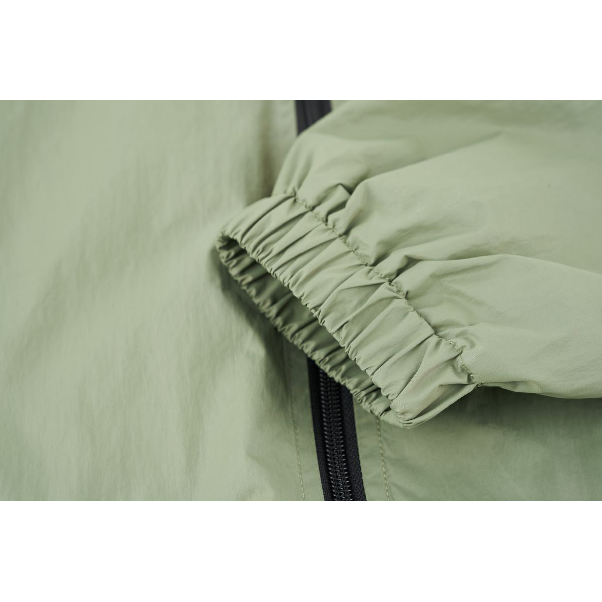 Close-up of a pale sage-green jacket sleeve with an elasticated cuff beside the black front zipper.