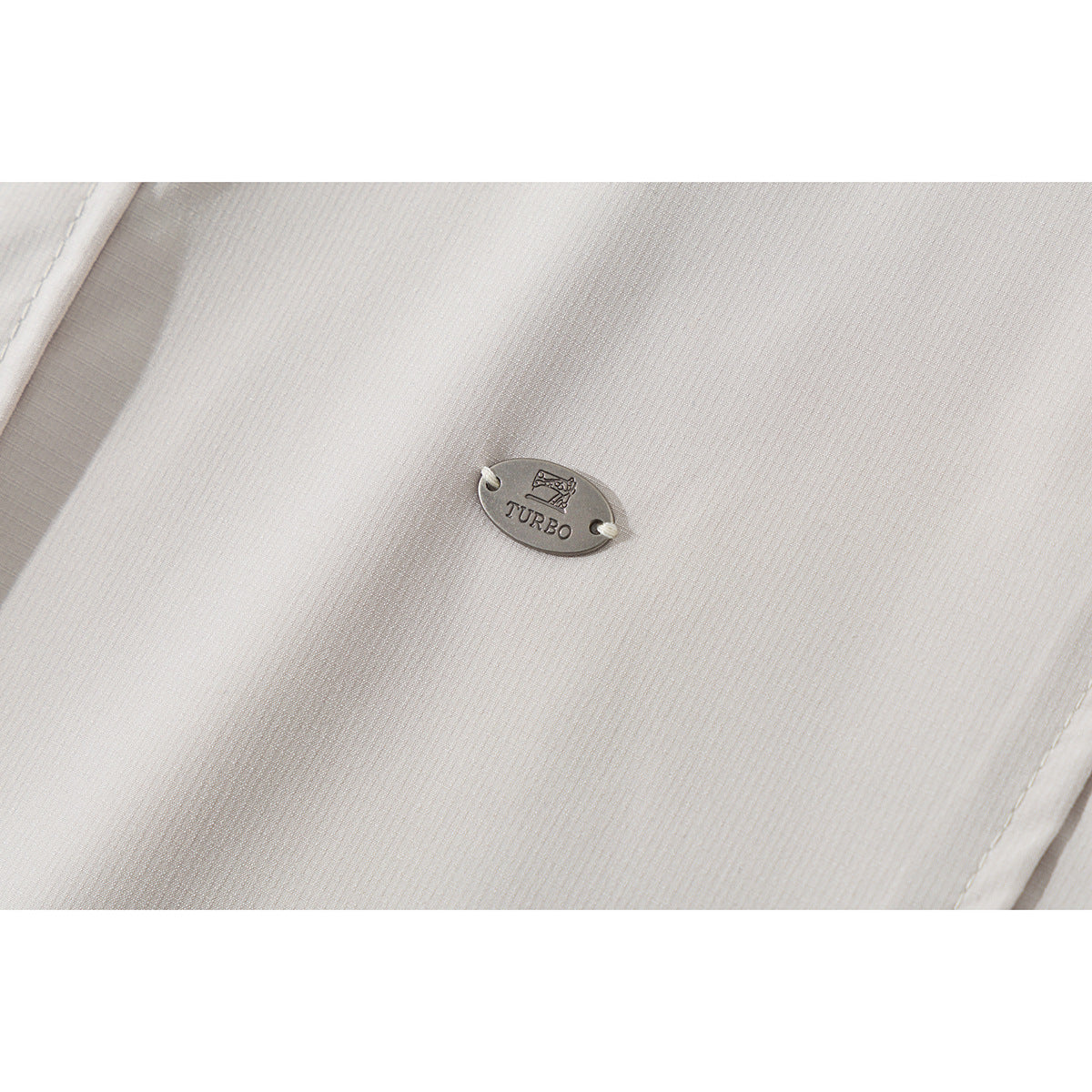 Close-up of a small oval TURBO metal tag attached to light beige shirt fabric.