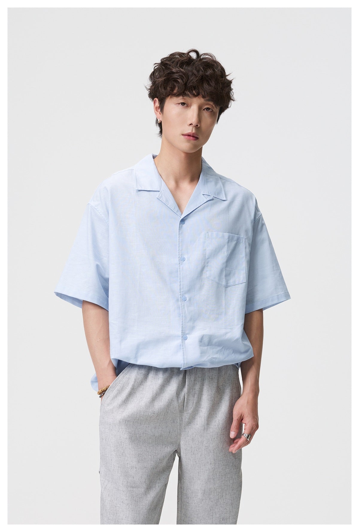 Model wearing a light blue short-sleeve button-up shirt with a notched collar and chest pocket, paired with gray trousers.