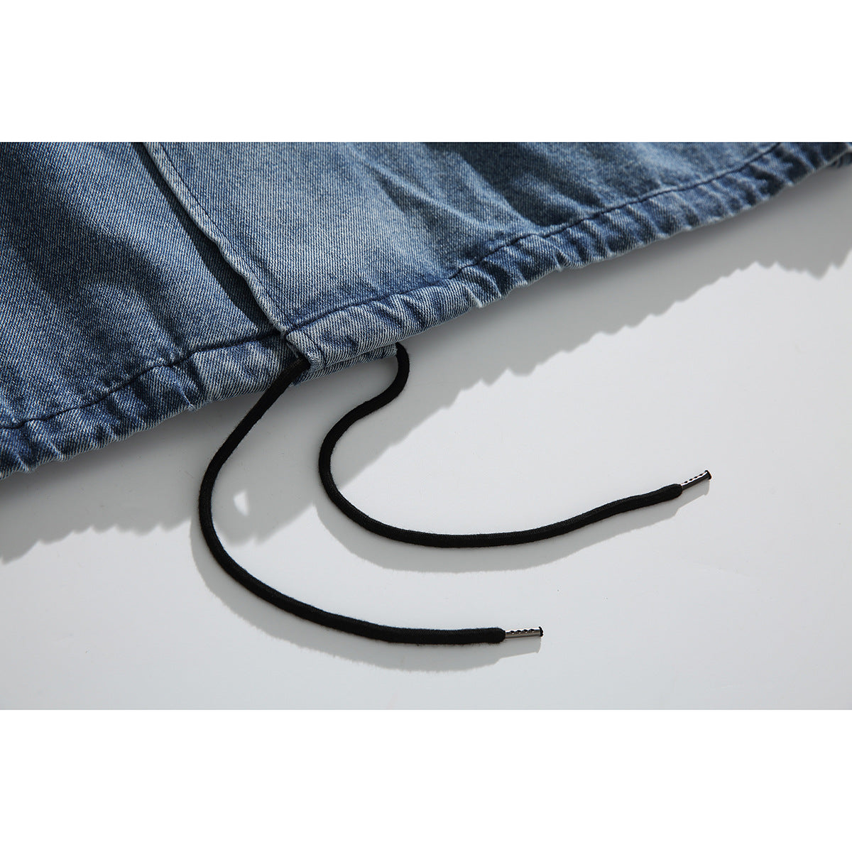 Close-up of the washed blue denim jacket hem with two black drawcords and metal tips.