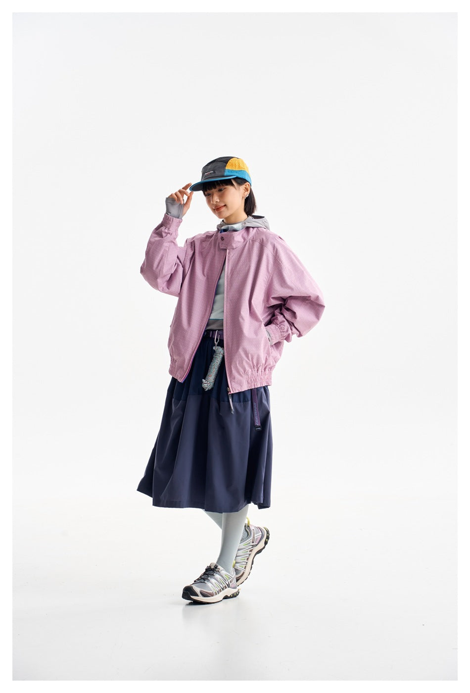 Model wearing a pink zip jacket with elastic cuffs and hem, paired with a navy skirt and color-block cap.