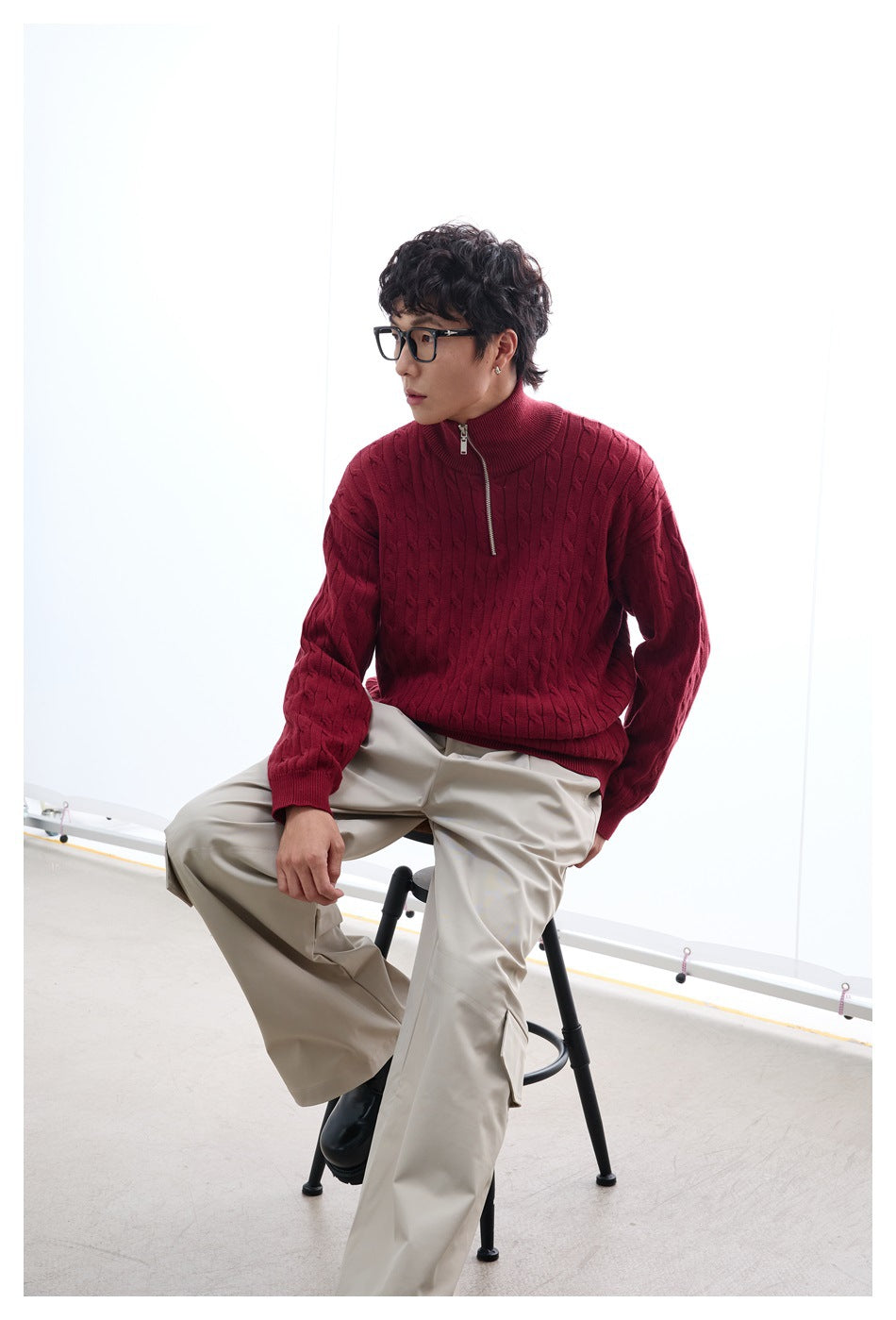 Man seated on a stool wearing a red cable-knit half-zip sweater with beige cargo pants