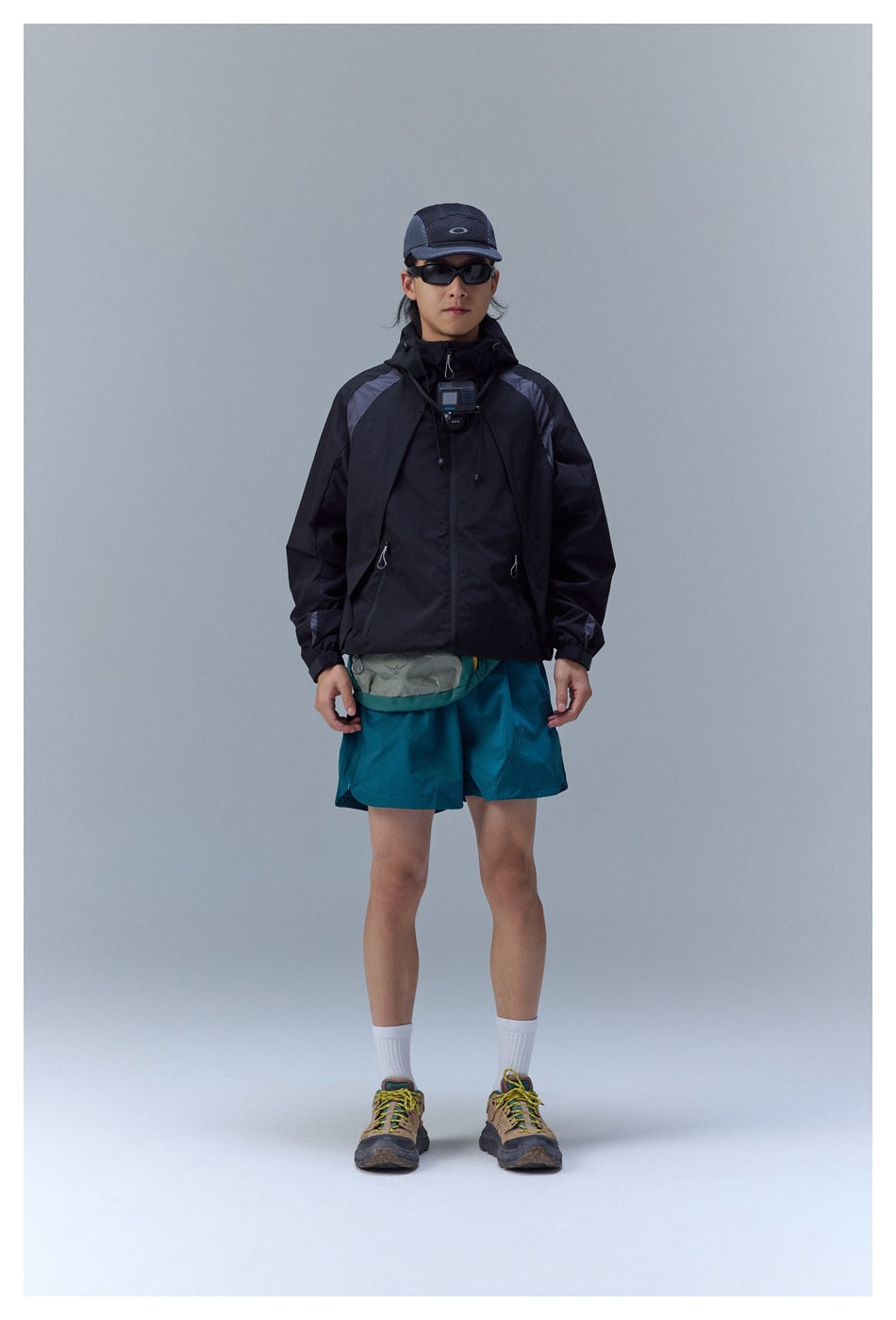 Front view of a model in a black hooded jacket with gray shoulder panels and teal shorts.