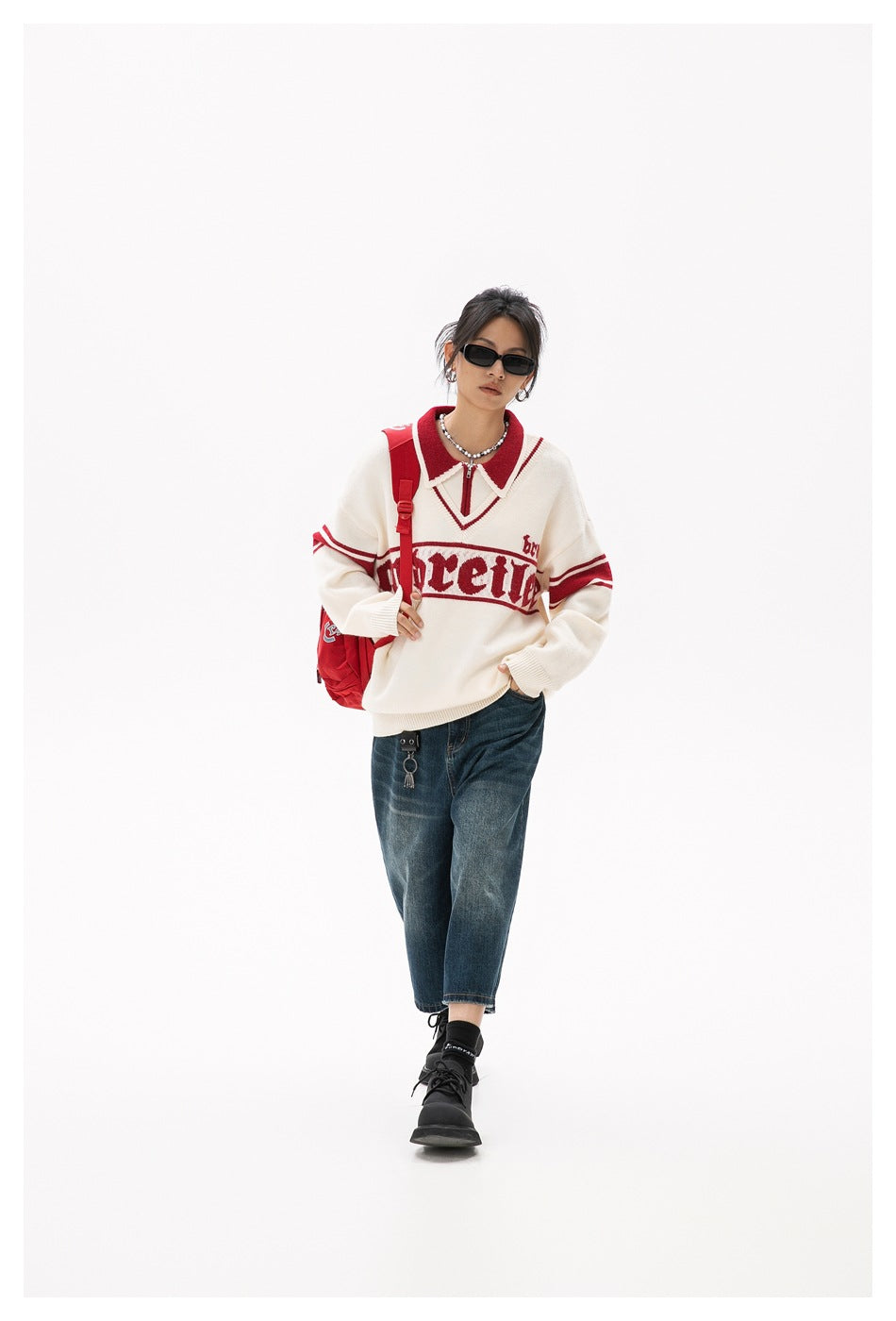Model wearing an ivory sweater with red collar, sleeve stripes and chest lettering, paired with dark jeans.