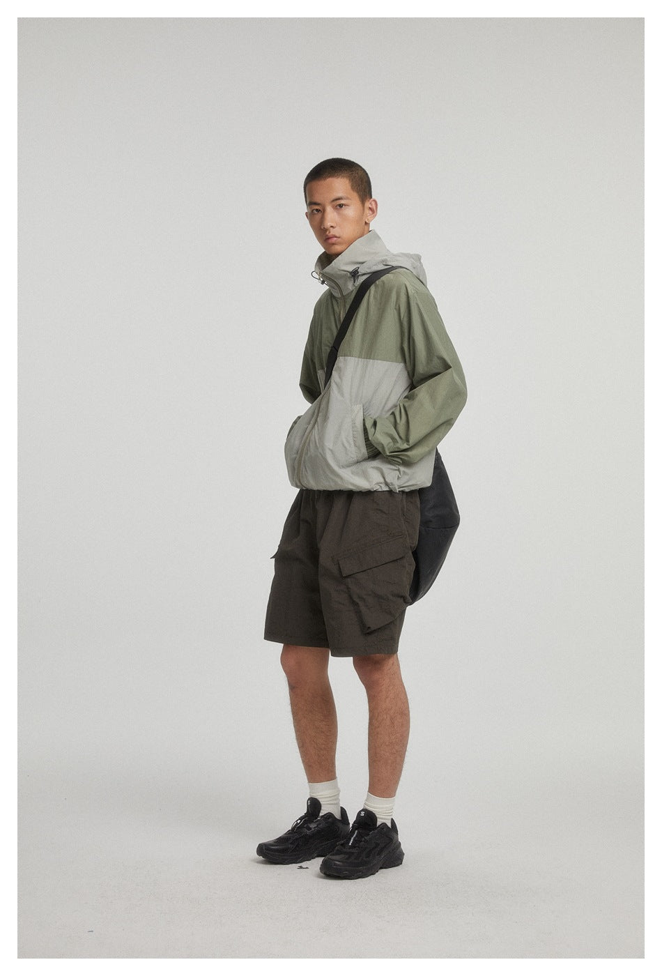 Three-quarter view of a model in an olive and light gray hooded jacket with dark cargo shorts and a black shoulder bag.