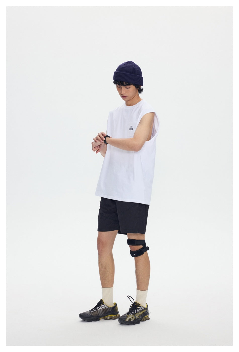 Model wearing a white sleeveless pocket tee and black shorts while checking a wristwatch.