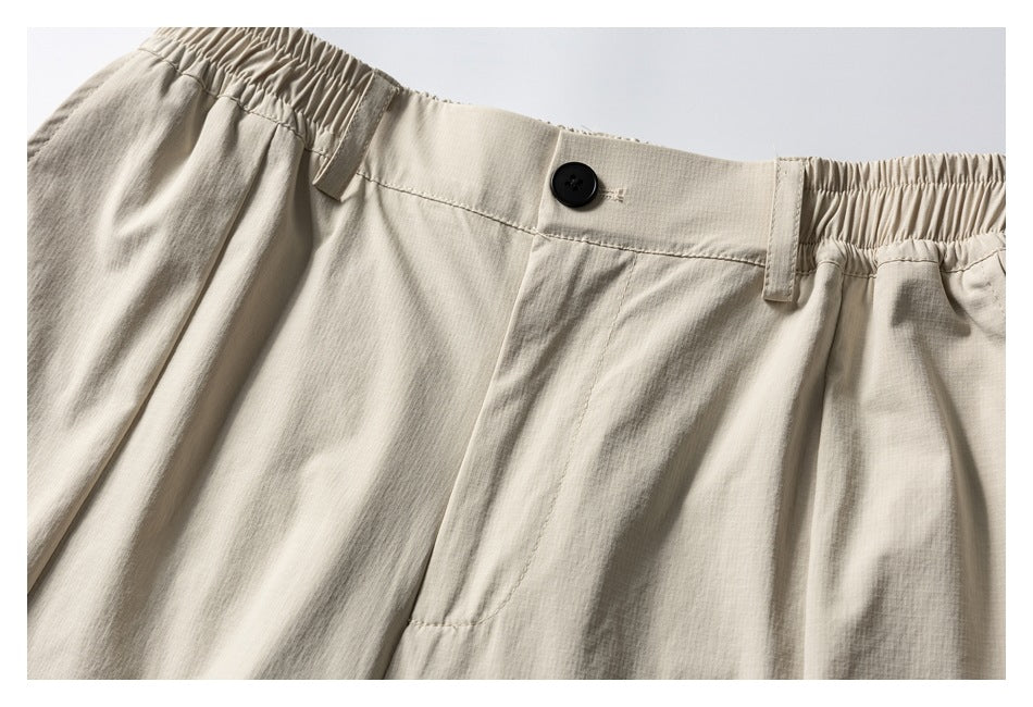 Close-up of cream trousers showing the waistband, black button, belt loops, elasticated sides and front pleats.