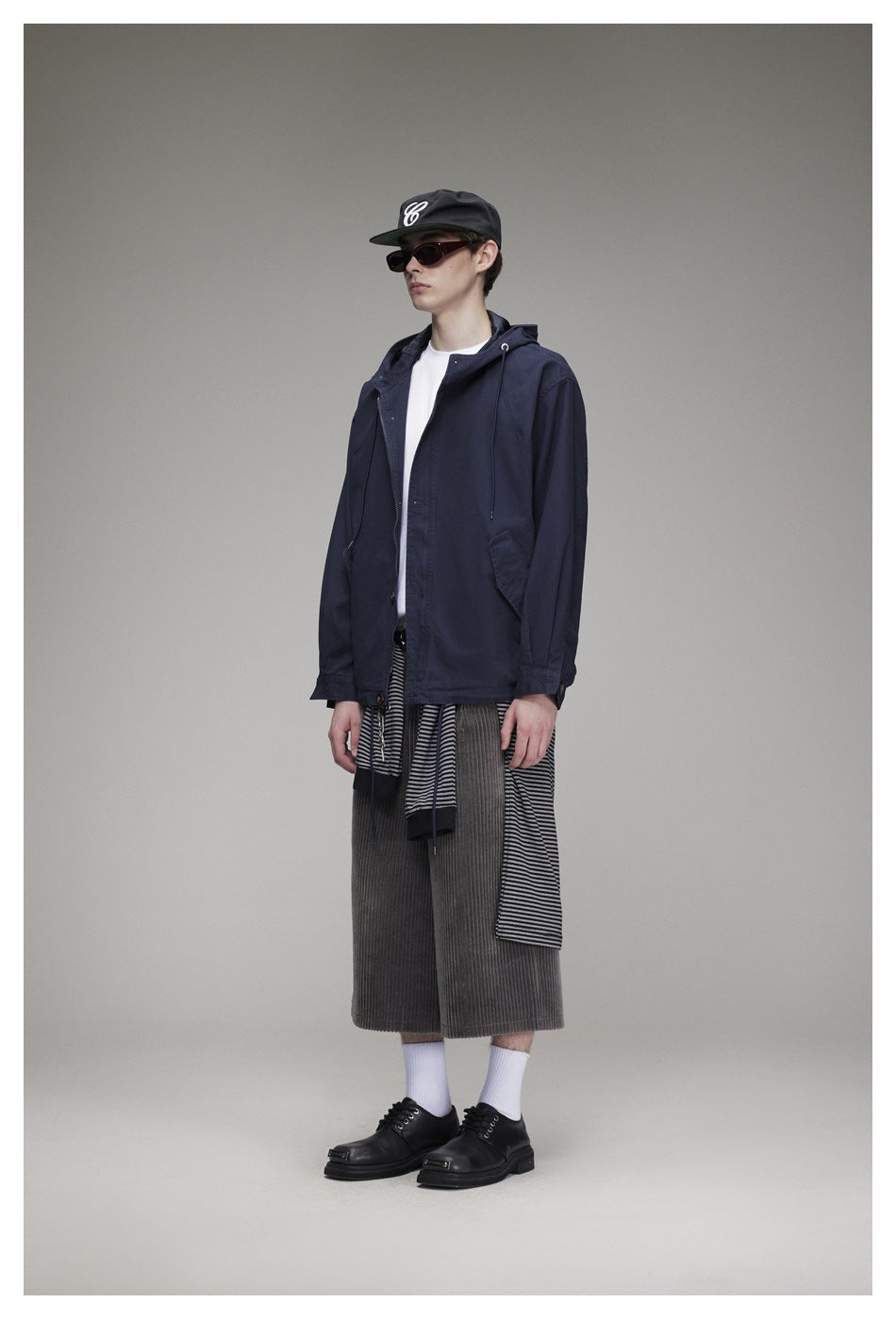 Model wearing a navy hooded jacket, white shirt, striped shorts, and black cap and shoes.