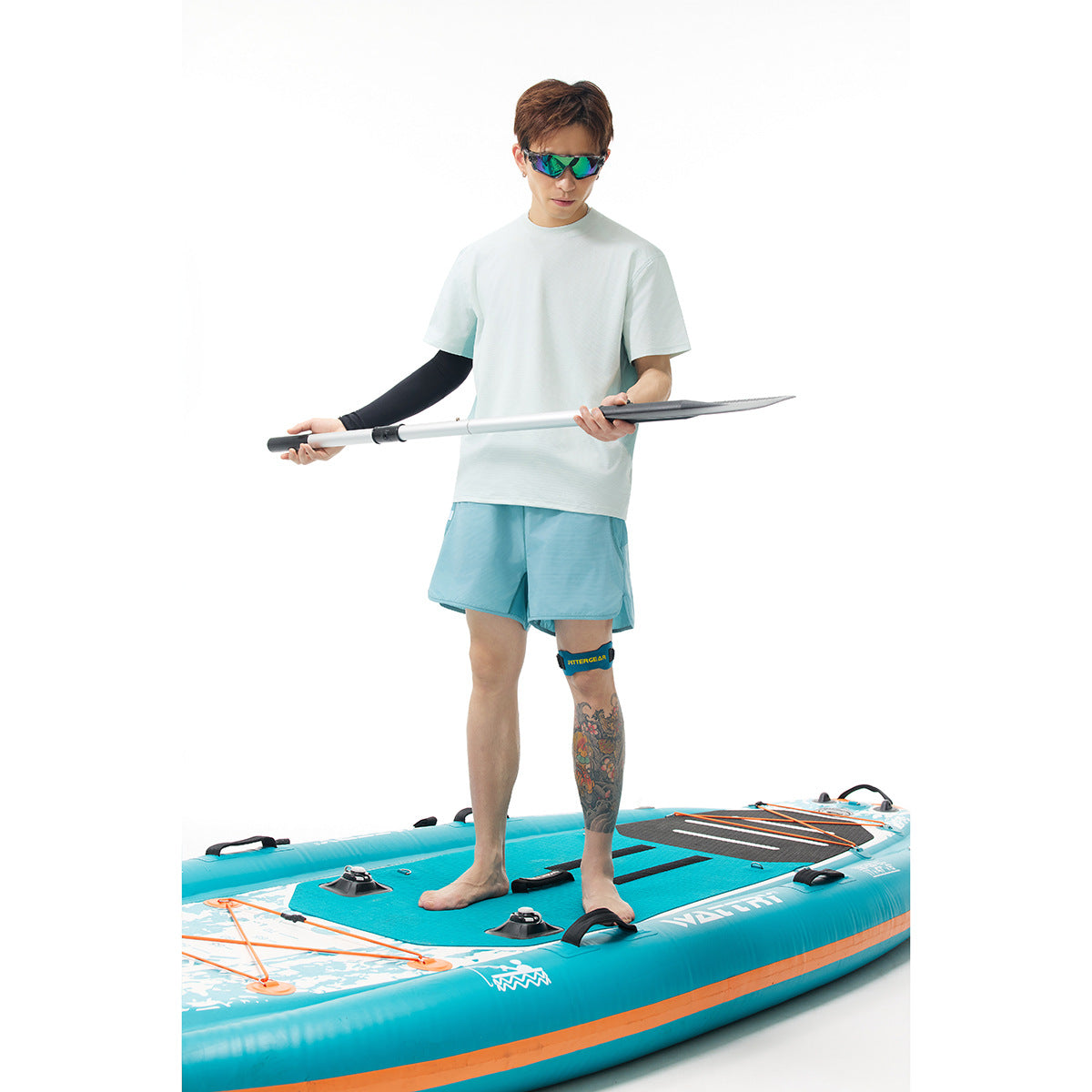 Model wearing a white T-shirt and aqua shorts while standing on a turquoise paddleboard with a paddle.