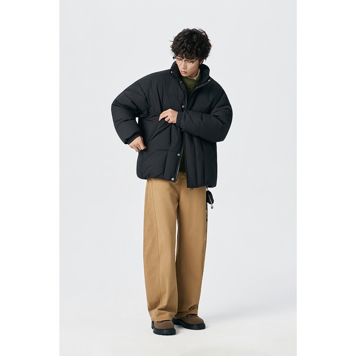 Model wearing a black padded jacket and tan trousers, adjusting the front closure.
