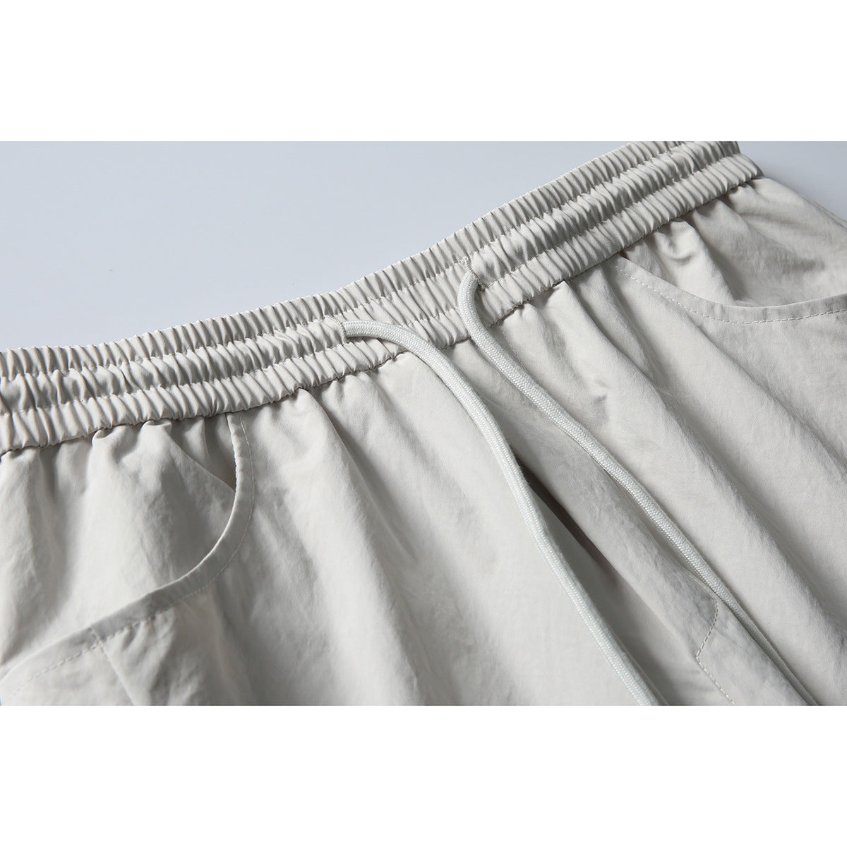Close-up of light gray pants showing the gathered elastic waistband, long drawstring, and front pocket opening.