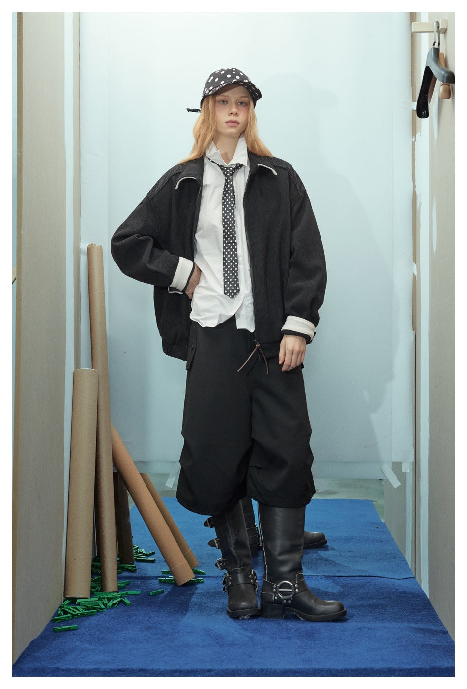 Full-length model in a black jacket with cream trim, white shirt, polka-dot tie and cap, wide black trousers and boots.