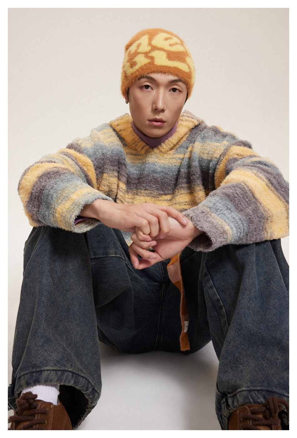 Seated model wearing a fuzzy hooded pullover with broad gray, mauve, and golden-yellow stripes.