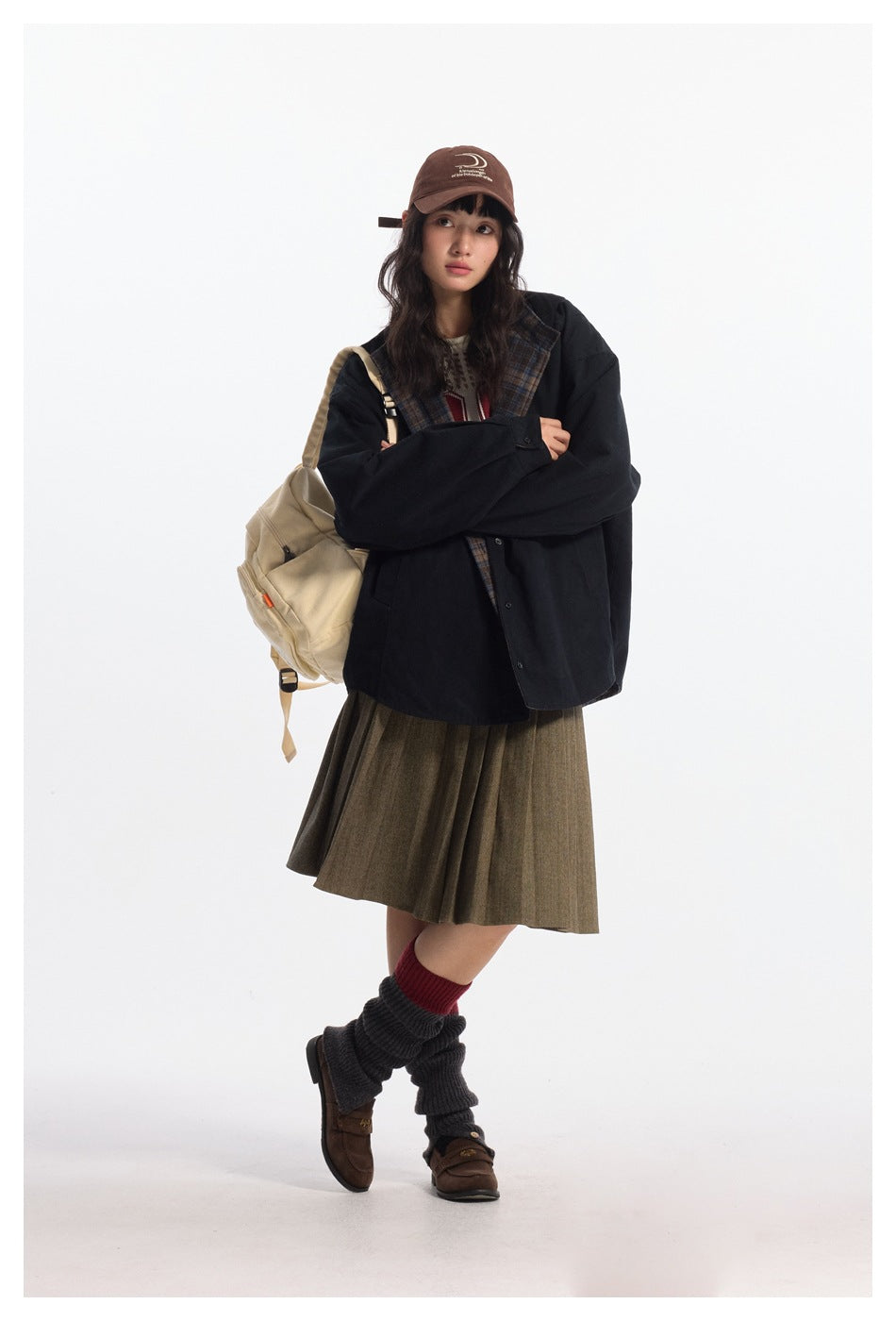Model in a black jacket with plaid lining, cream mesh top, olive pleated skirt, and beige backpack.