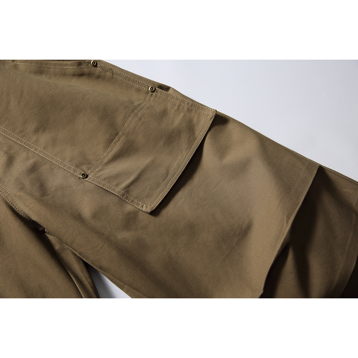 Close-up of olive-brown trousers showing a large stitched patch pocket with metal rivets.